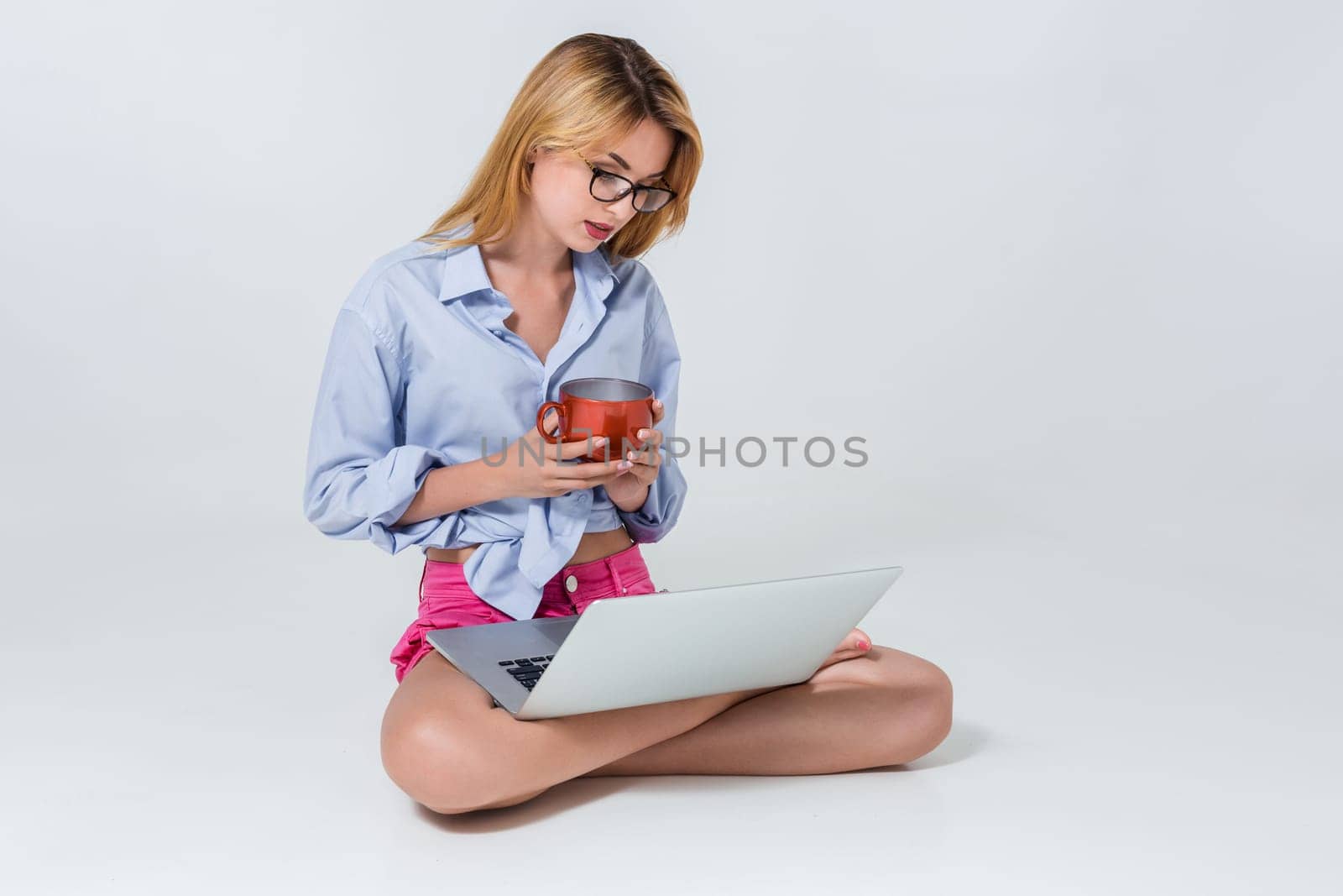 young woman sitting on the floor with crossed legs and using laptop on white background. satisfied, pleased, happy, smiling and looking at the monitor, holding a cup in his hands