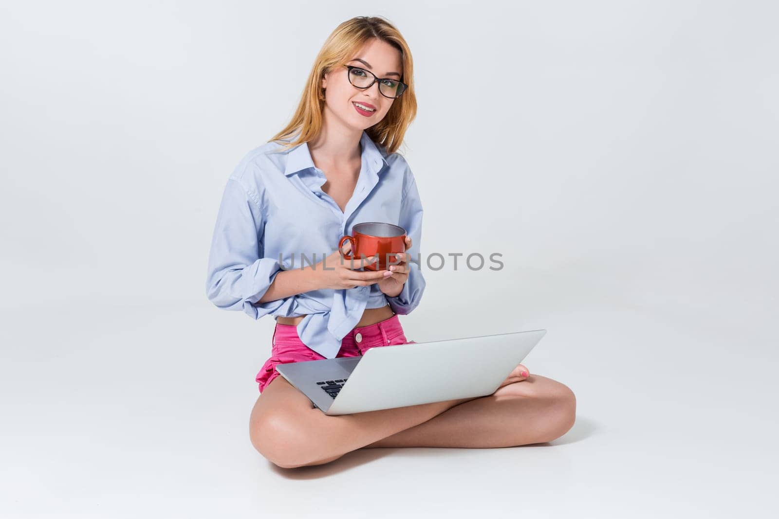 young woman sitting on the floor with crossed legs and using laptop on white background. satisfied, pleased, happy, smiling and looking at the camera, holding a cup in his hands
