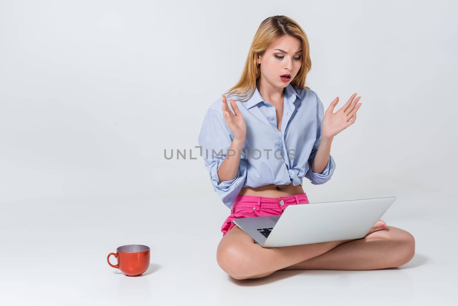 young woman sitting on the floor with crossed legs and using laptop on white background. Surprised, emotional