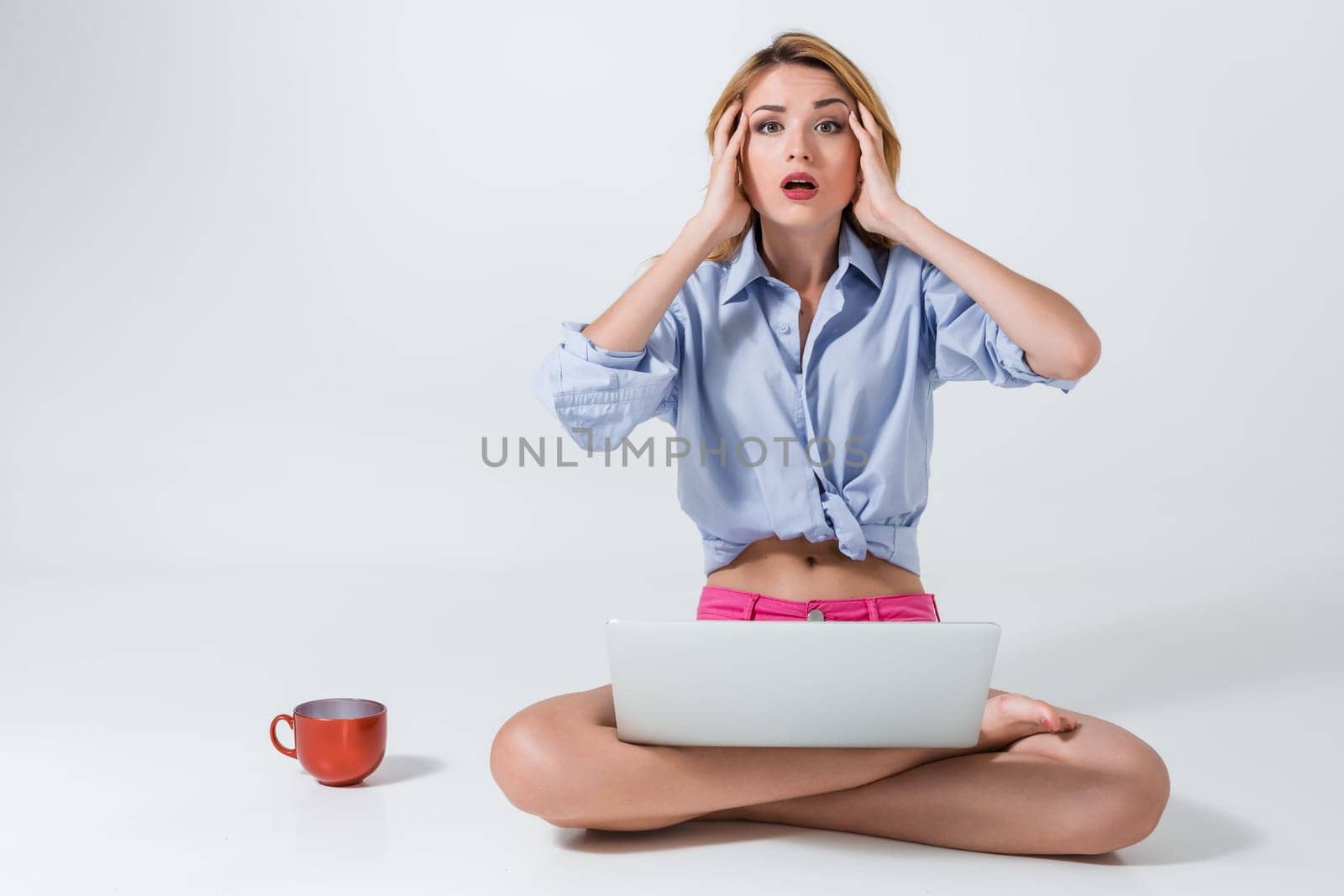 young woman sitting on the floor with crossed legs and using laptop on white background. tired, surprised, tortured, his hands holding his head