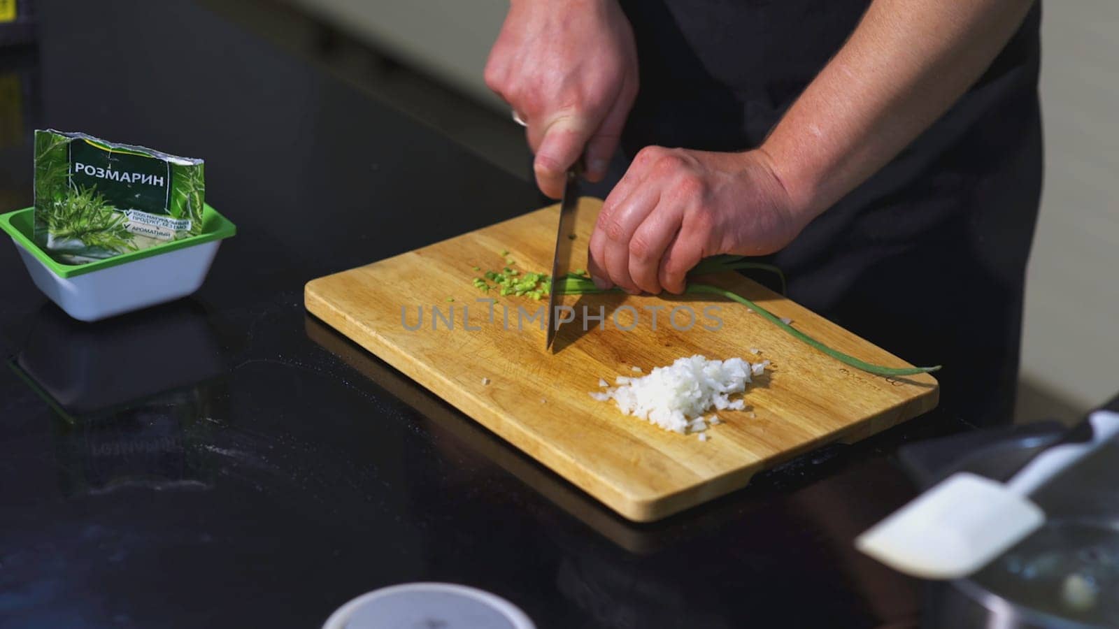The cook is cutting green onions. ART. Male hands in the frame are cutting green onions with knife on the board. On the table are board, onion, rosemary and spatula.