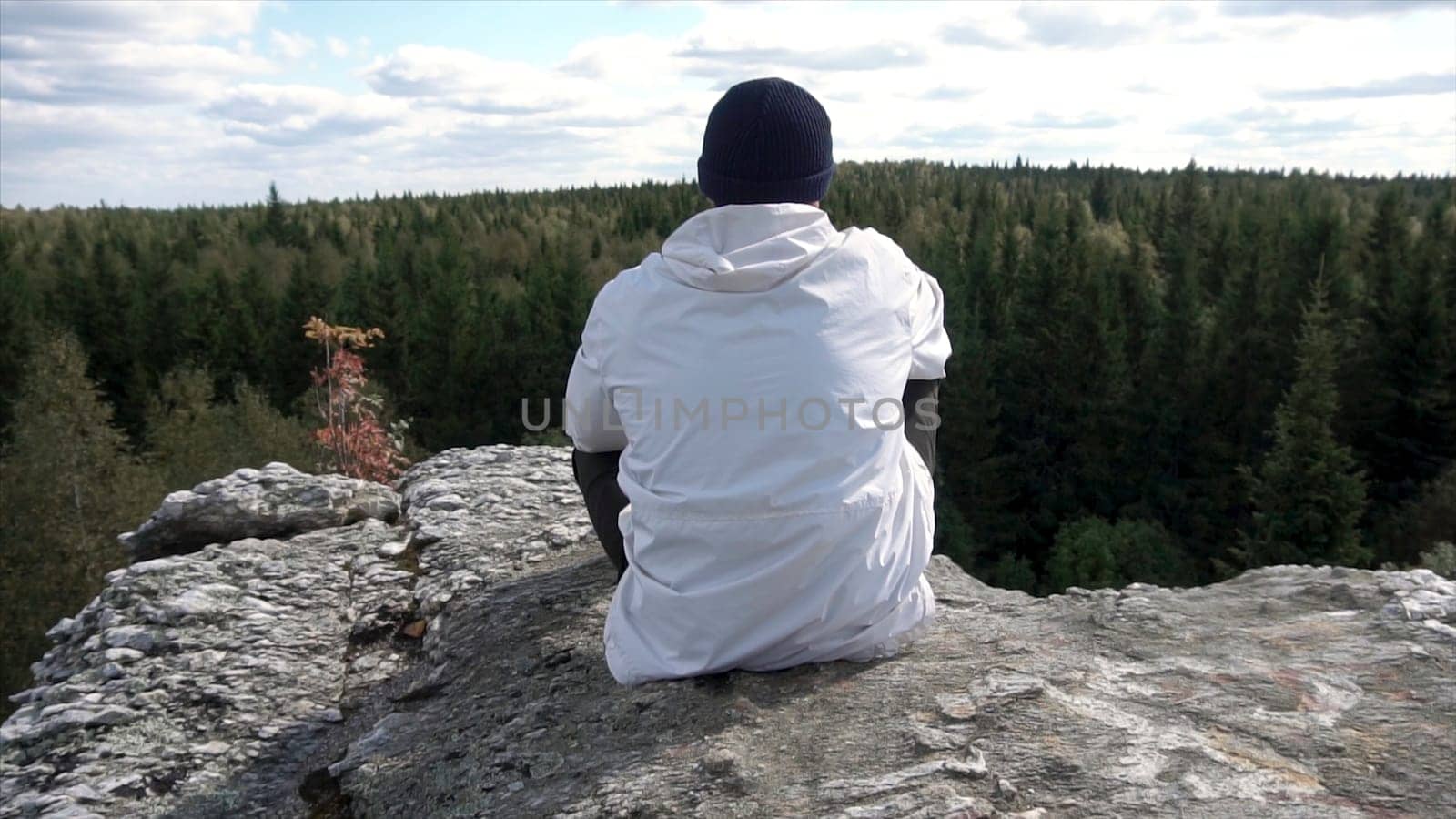 Alone man sitting on a high mountain top looking at a cloudy sky and trees peaks. Rear view of a man in the hat and white jacket sitting on a rock, looking at beautiful forest