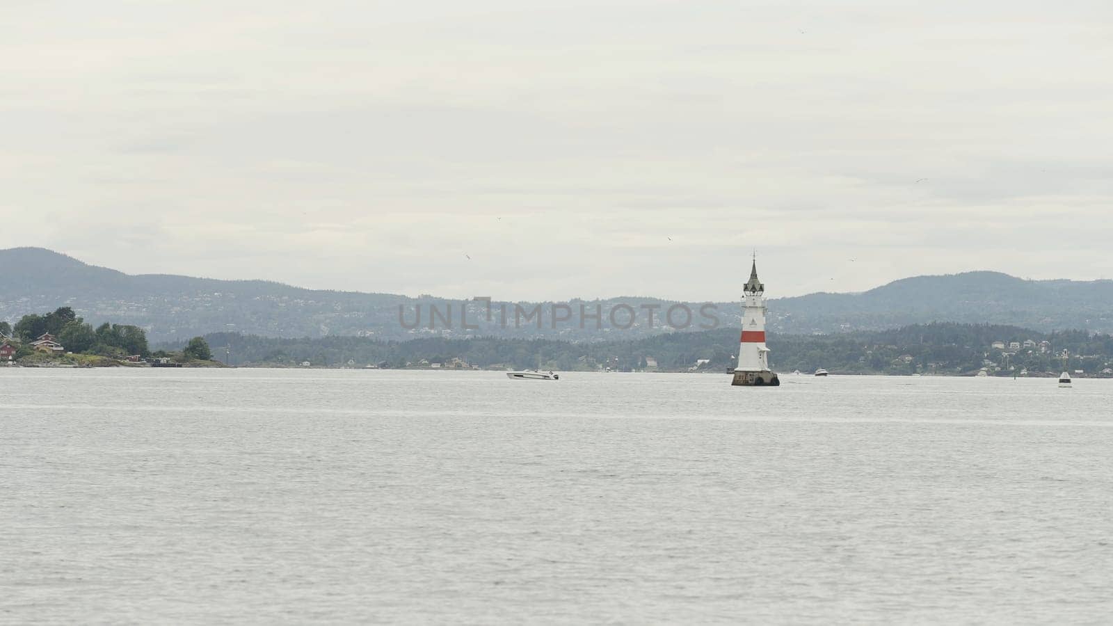 Picturesque natural landscape with a lighthouse and rippling water surface. Action. Cloudy sky above ocean and mountains on the background