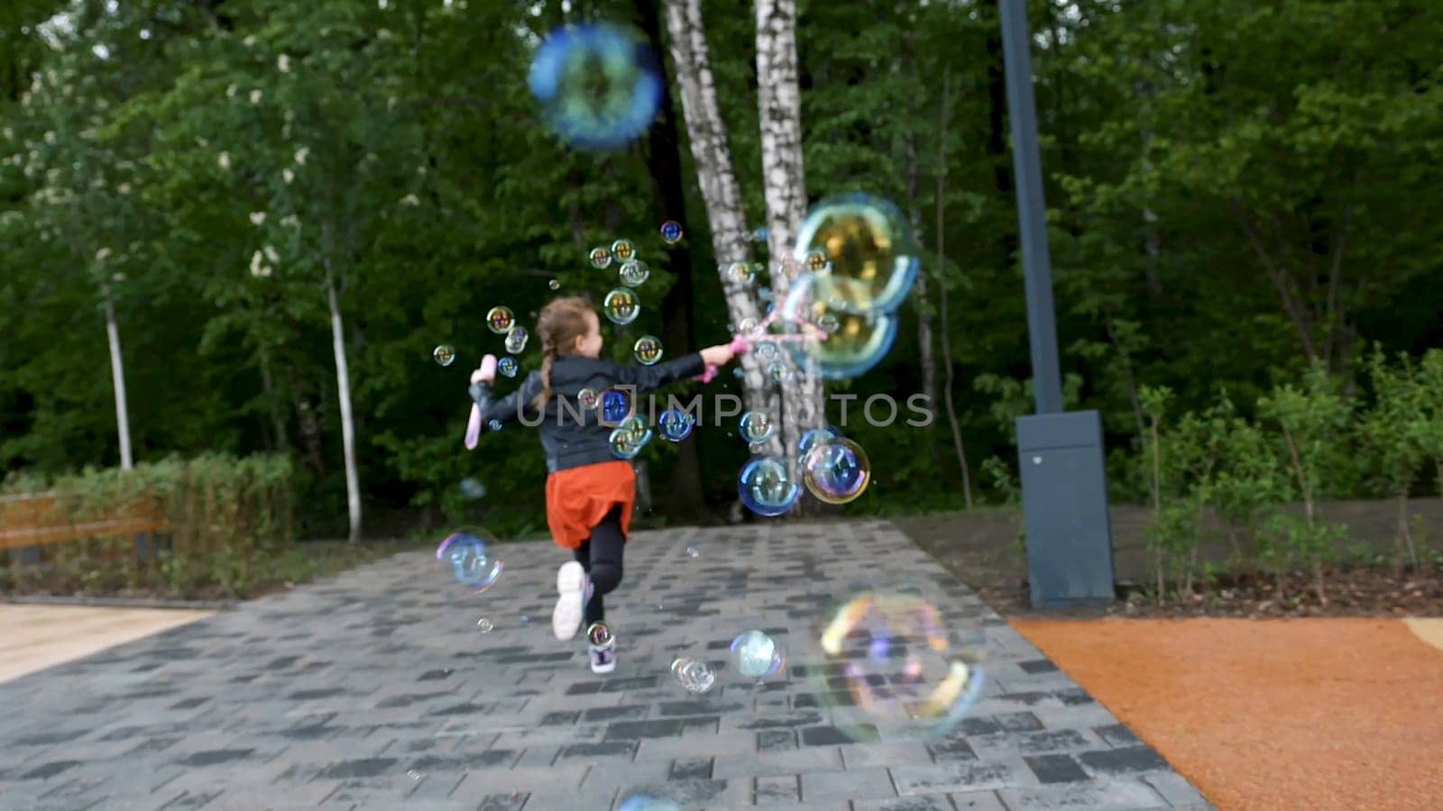 Lovely girl blowing bubbles while running, rear view. Female caucasian child dressed in warm clothes creating many soap bubbles in spring time on the background of green trees of a city park.