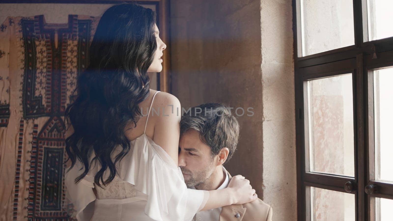 Elegant couple at window in old house. Action. Couple of lovers hug at window. Beautiful couple hugging looking out window by Mediawhalestock