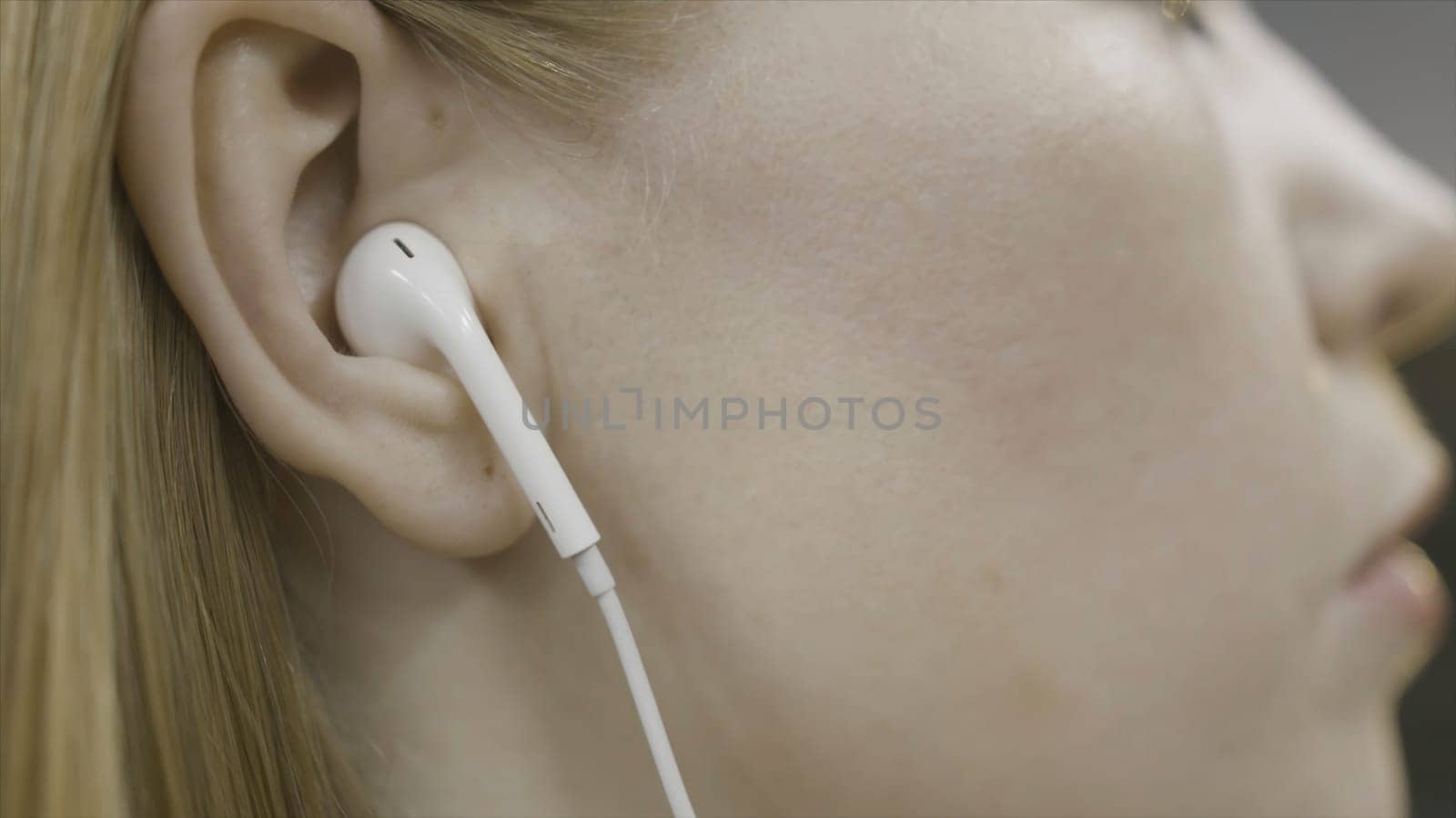 Close up side view of blonde woman head wearing headphones and touching her hair. Girl with white earphones tucking her hair behind her ear.