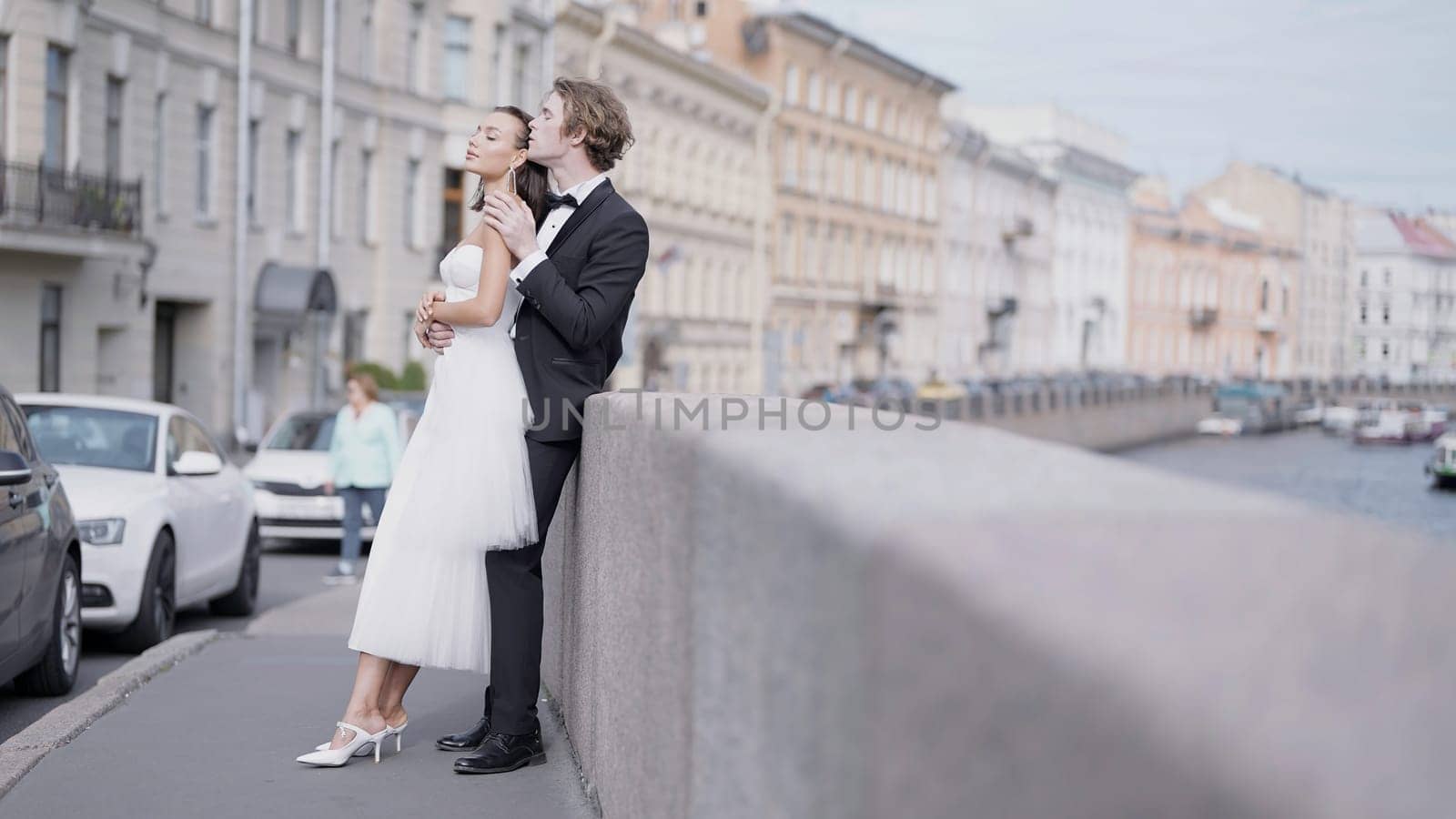 Side view of caring lovely just married couple embracing by the granite sidewalk. Action. Elegant bride and groom outdoors in the street