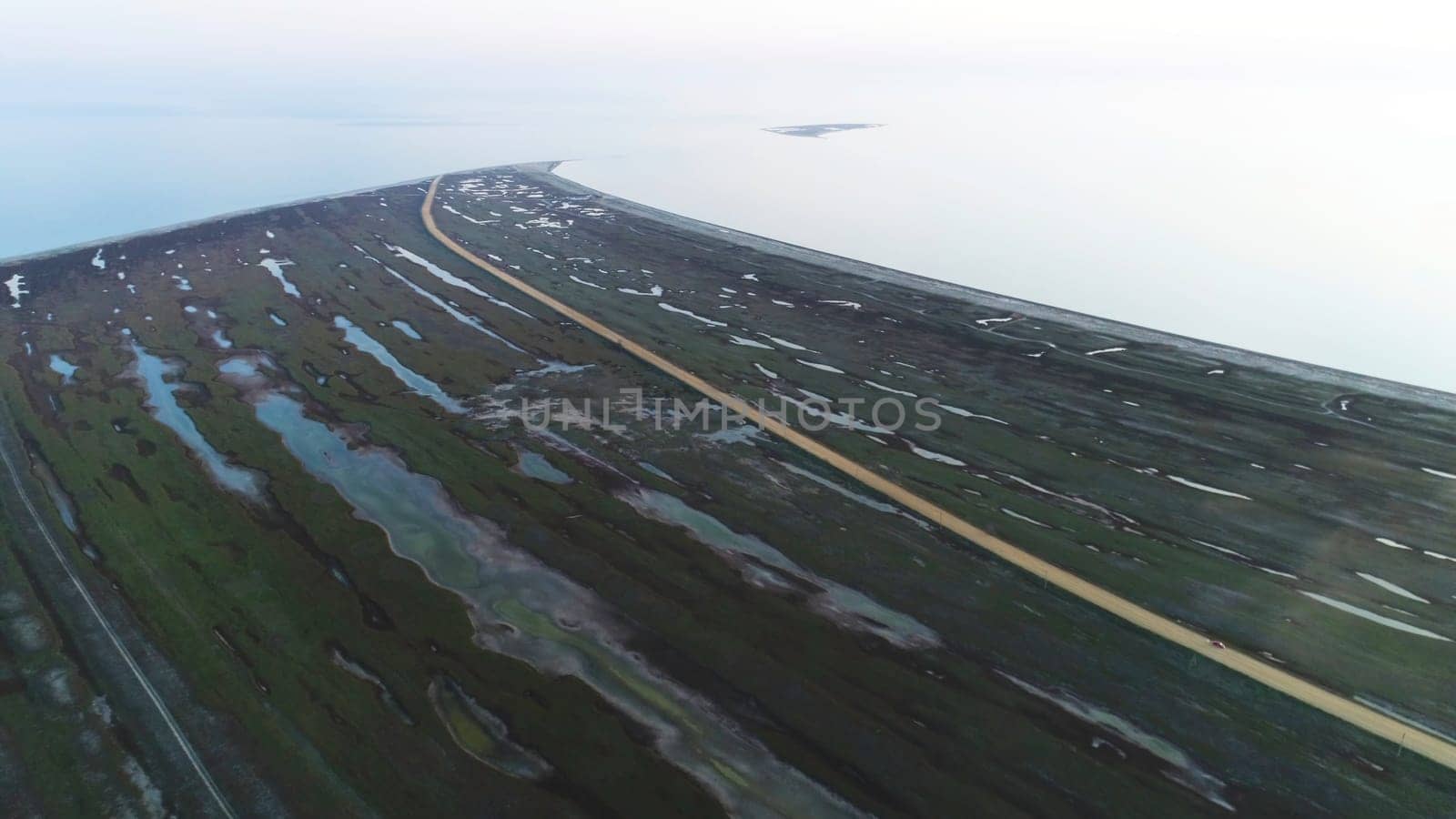 From above view of a meadow field near the sea shore and a yellow country road. Aerial for a abandoned flooded green field in a cloudy day.
