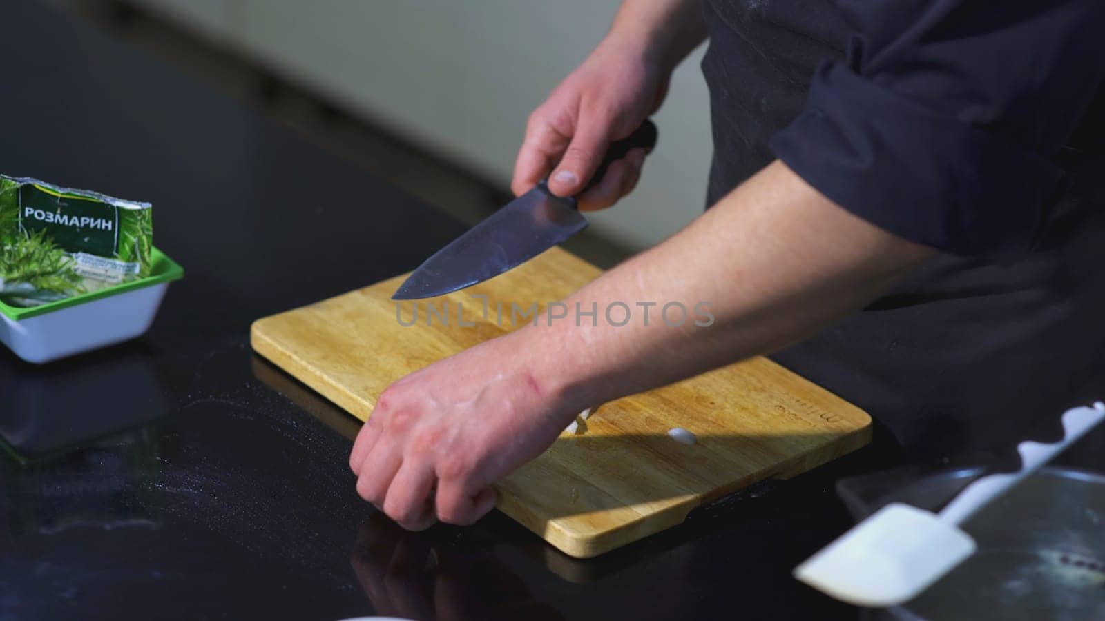 The cook cuts the onion. ART. Man in the kitchen cuts white onion into half rings with knife. Next to the board is spatula and bag of rosemary.