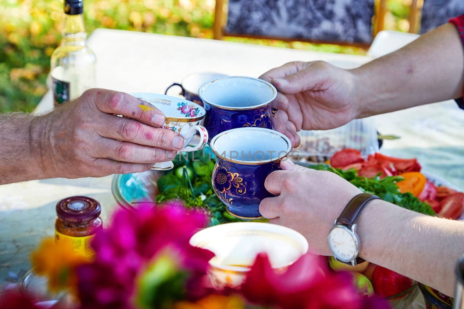 People's hands with beautiful cups of tea at the outdoor table on a sunny summer day. A joint tea party in the landscape, a picnic