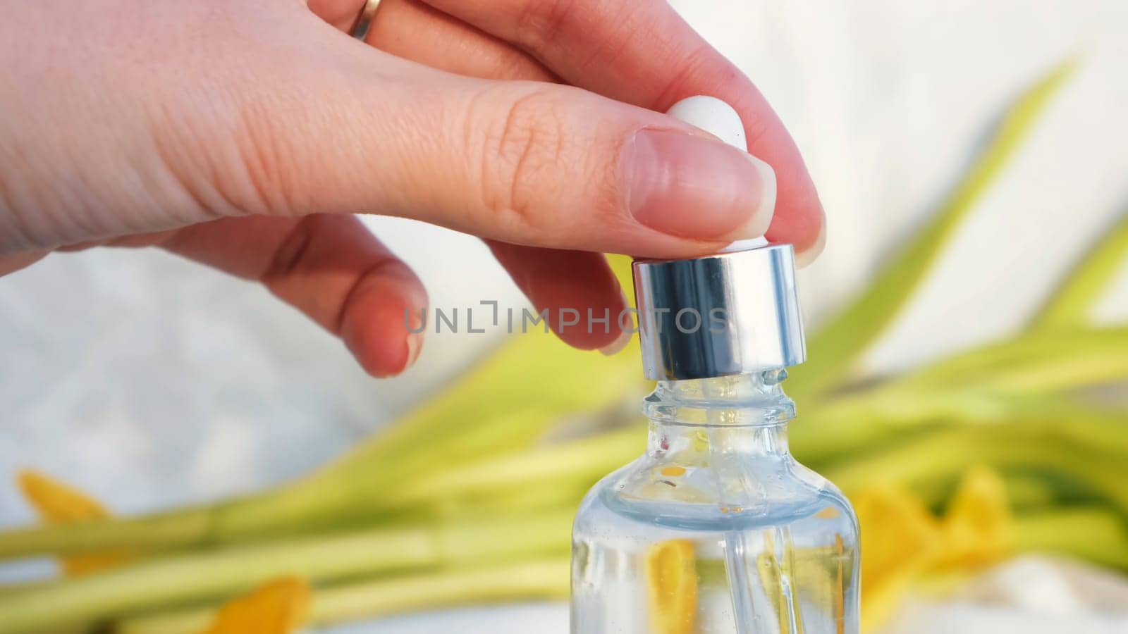 Vial with essential oil and yellow soft petals isolated on a white textile background. Girl hand touching cosmetic package surrounded by tulip leaves.