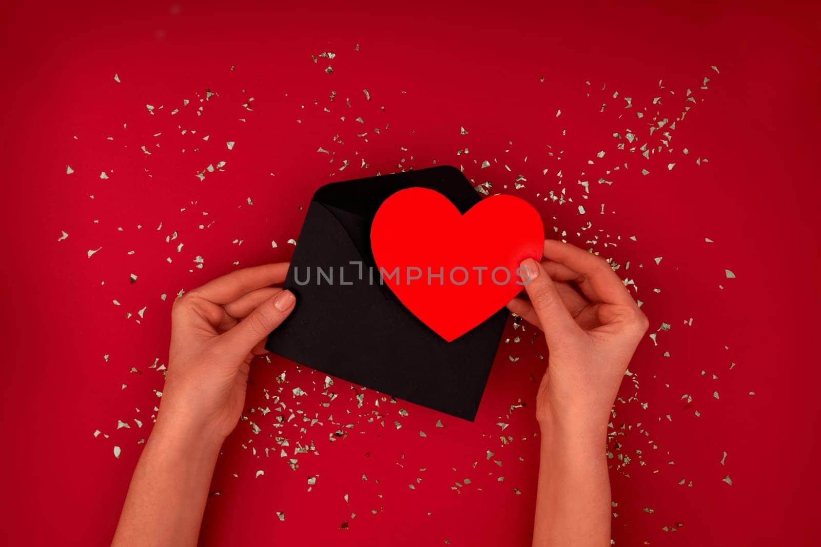 Female hands takes out and receives or sends a red card in the shape of heart from envelope on red maroon background with scattering of gold sparkles and glitter, top view copy space