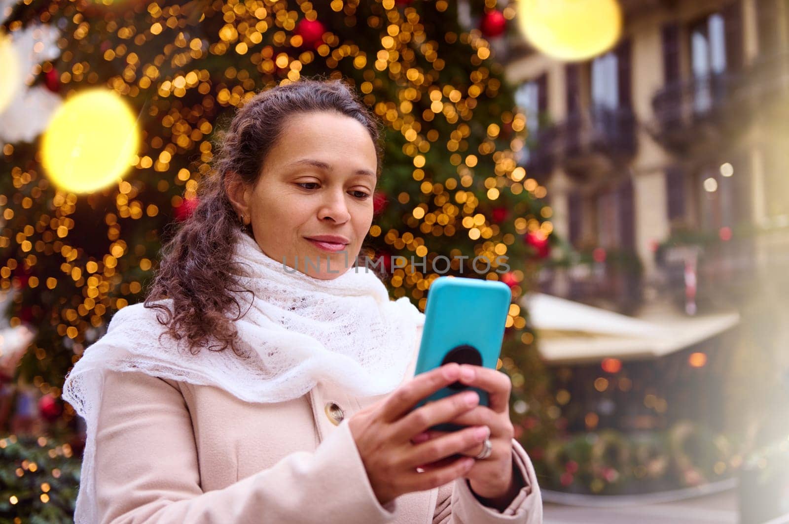 Closeup portrait of a multi ethnic beautiful woman holding mobile phone against illuminated Christmas tree with bokeh by artgf