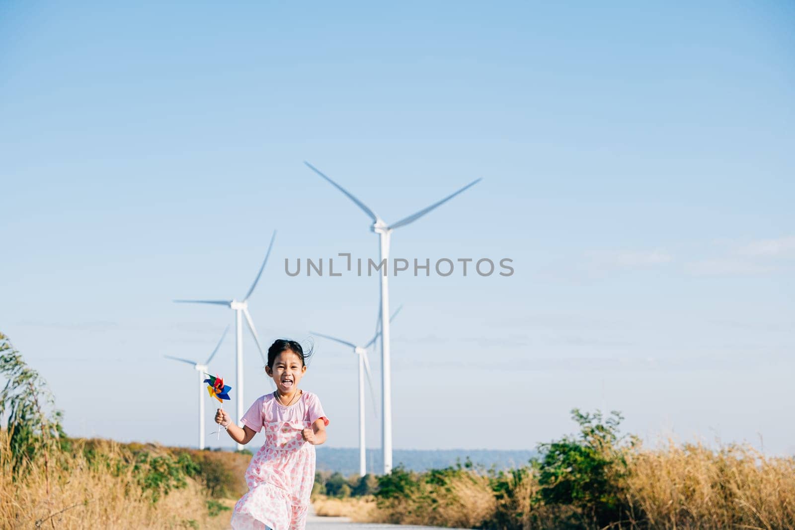 A cheerful girl runs near windmills holding pinwheels with joy. Embracing playful wind energy education in a picturesque wind turbine setting under a clear sunny sky.