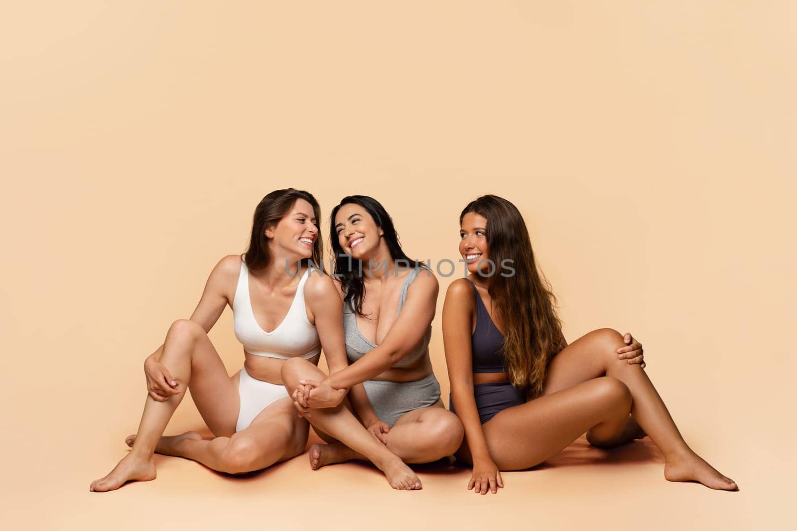 Three glad diverse millennial women in undergarments sit on floor, enjoy body positivity, beauty care, have fun, isolated on light studio background, full length. Friendship, confidence