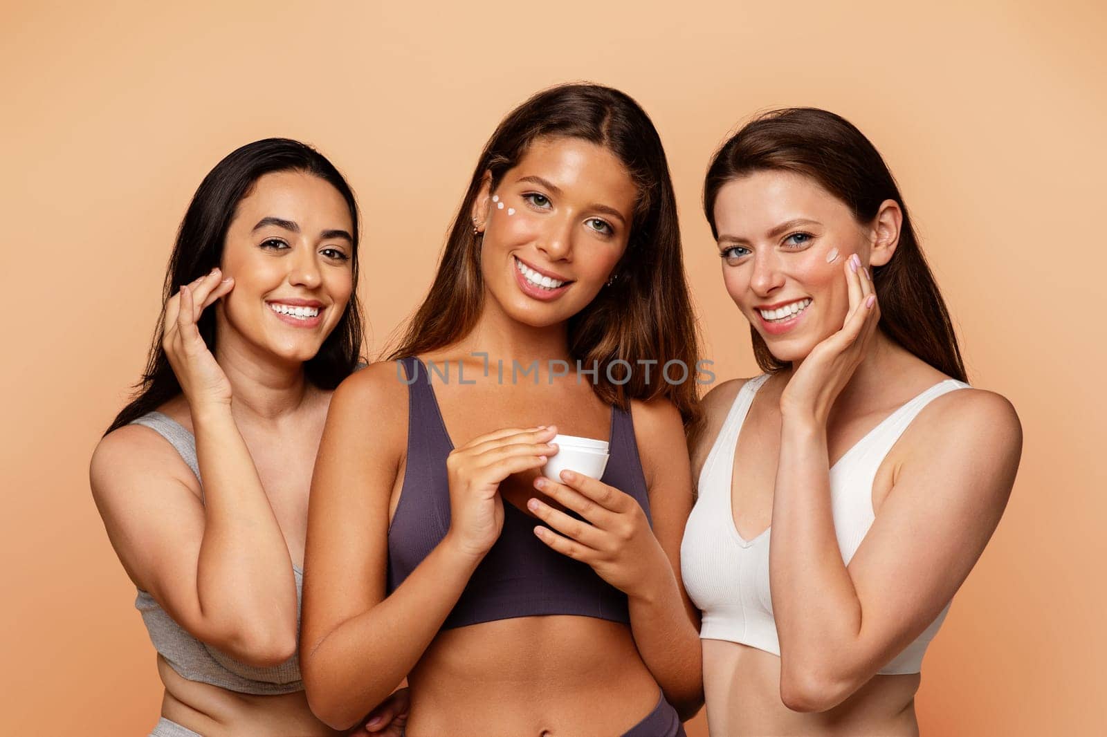 Three multiethnic young women in underwear show jar, put cream on face, enjoy beauty care, hydration at home on beige studio background. Fun lifestyle, cosmetics and spa, close up