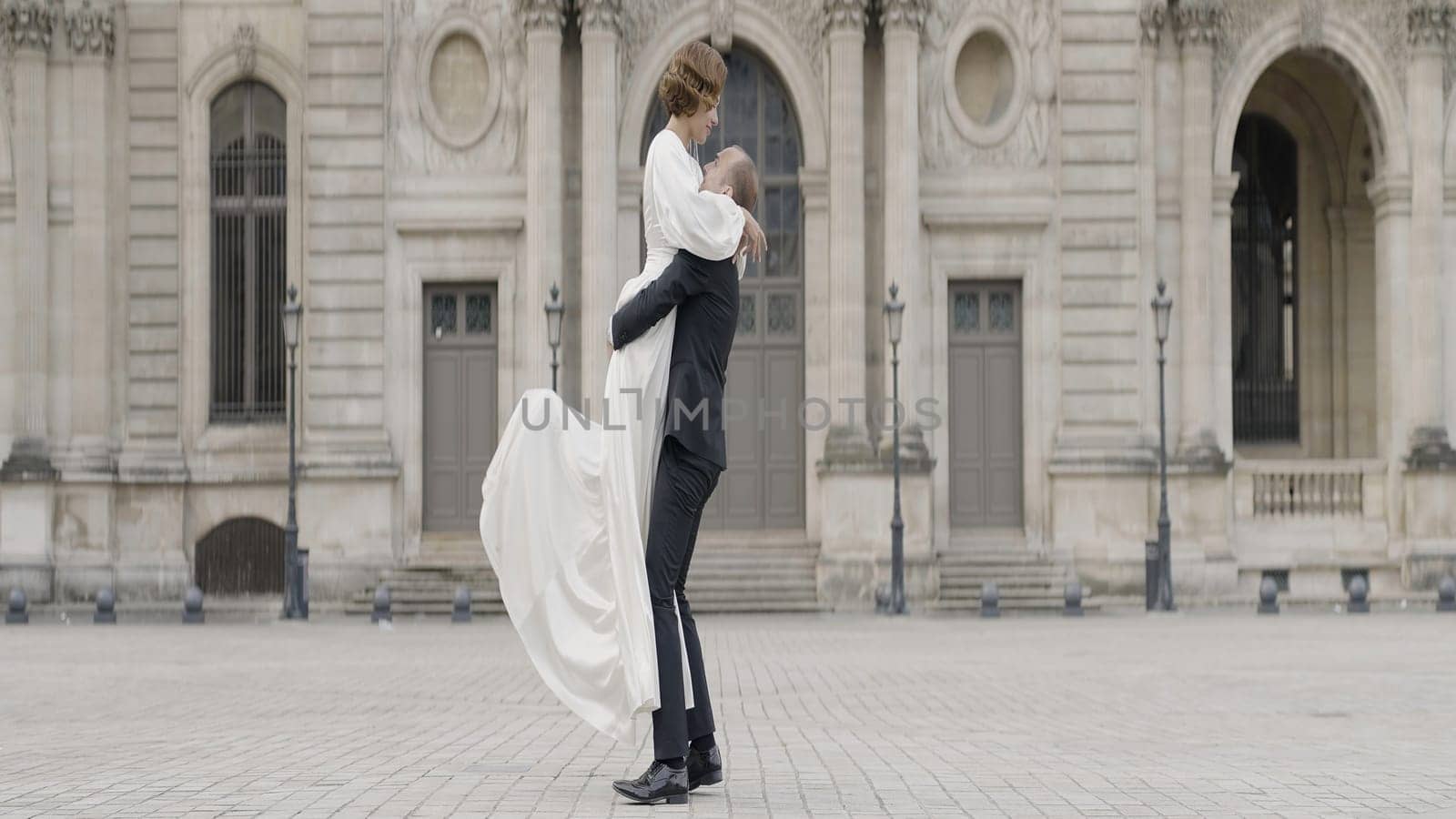 A man picks up his wife in his arms. Action. A date on the street where a girl in a white dress with a beautiful styling who jumps on her hands on a man in a suit in the fresh air. by Mediawhalestock