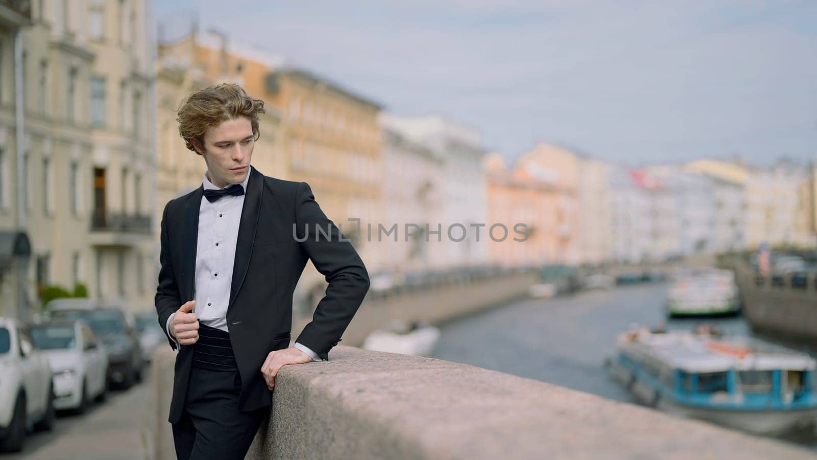 Models posing on the embankment . Action . A handsome, spectacular man in a suit with a bow tie and long hair standing with a young bride and posing for the camera. by Mediawhalestock