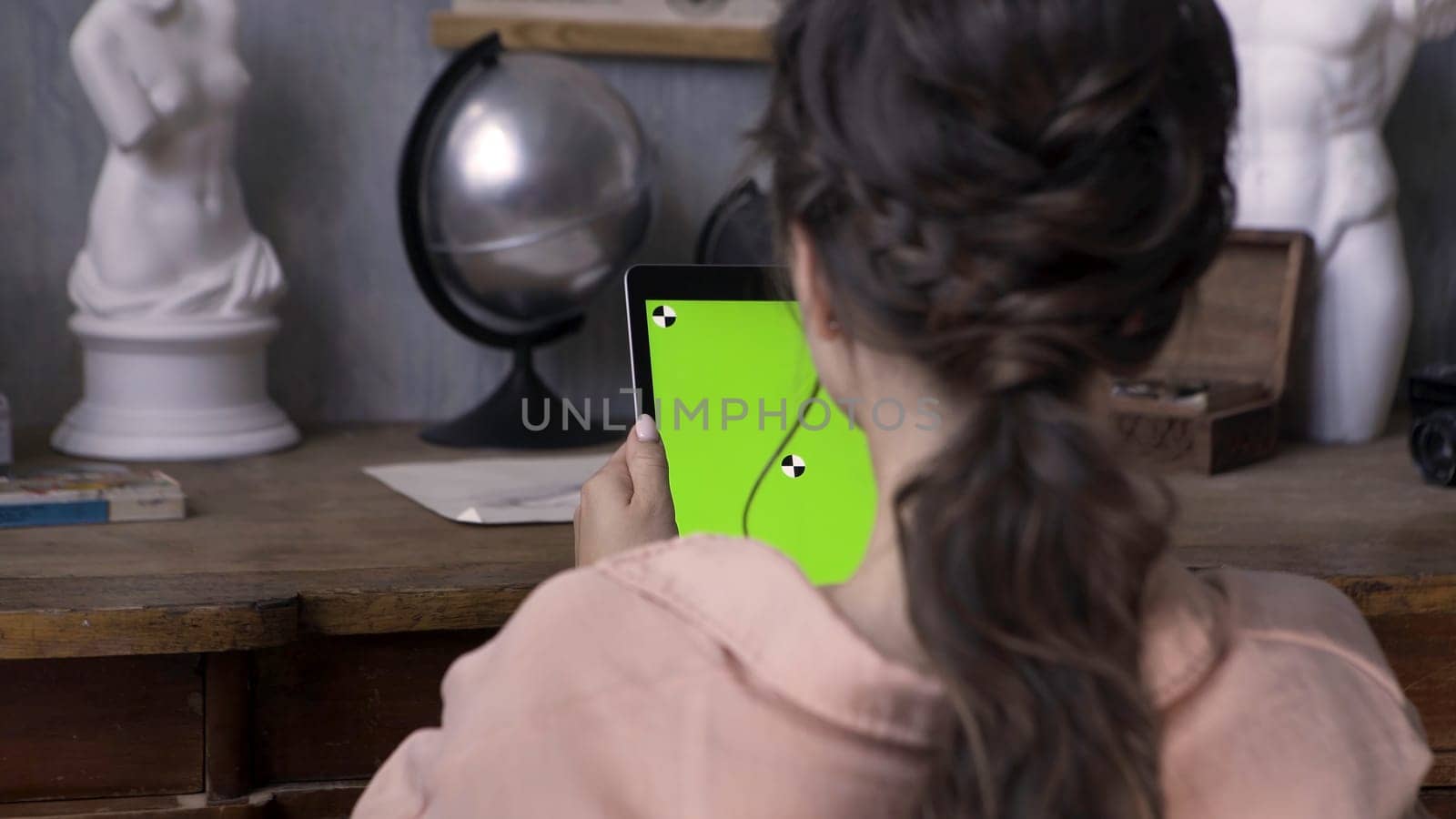 Woman hands holding tablet pc with green screen in the art studio with concrete wall and silver globe, plaster woman sculpture on the wooden table. Girl tapping on chromakey tablet.