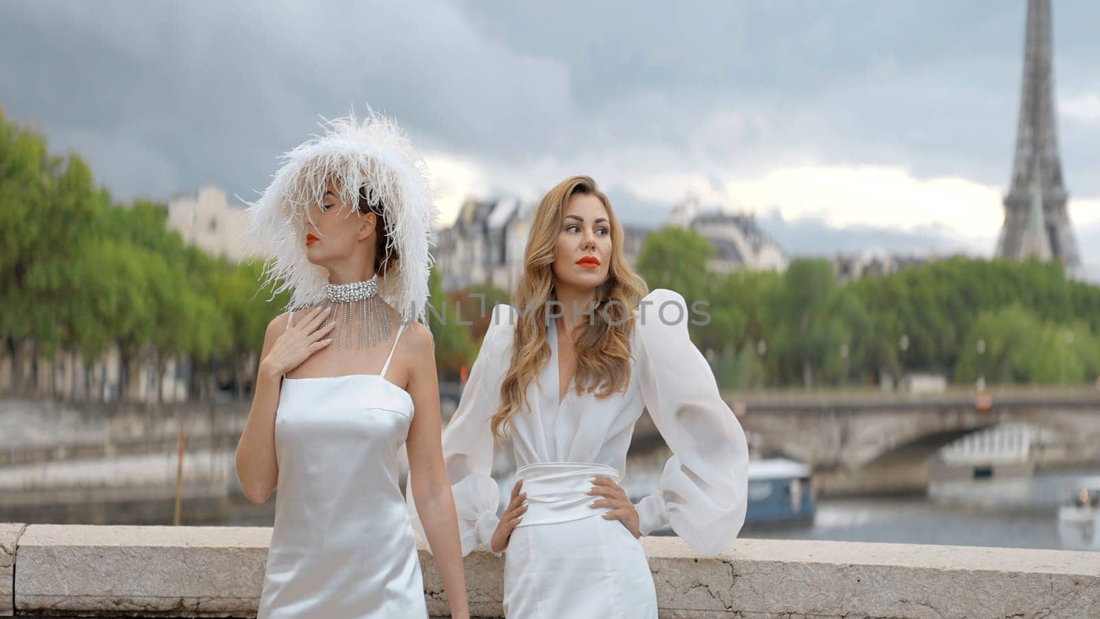 Stylish models in white dresses posing on the background of Paris streets and Effel tower. Action. Women in elegant dresses outdoors, one of them wearing a hat with feathers