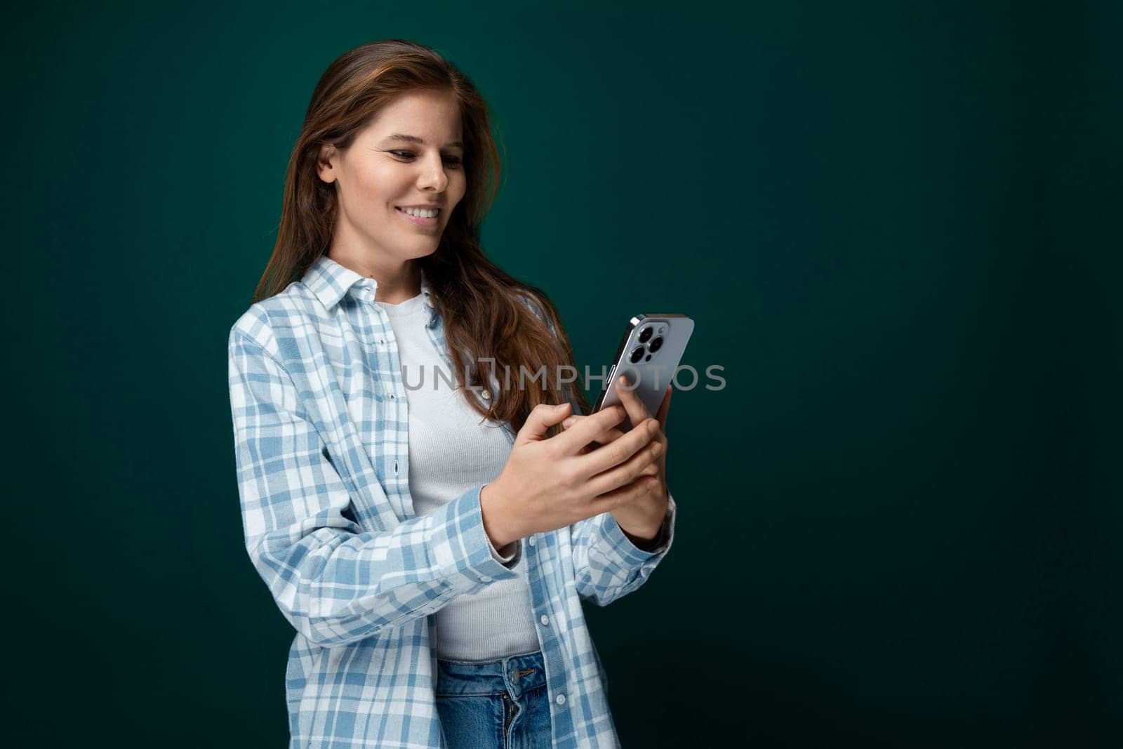 A pretty young woman with brown eyes dressed in a checkered blue shirt is chatting on a smartphone.