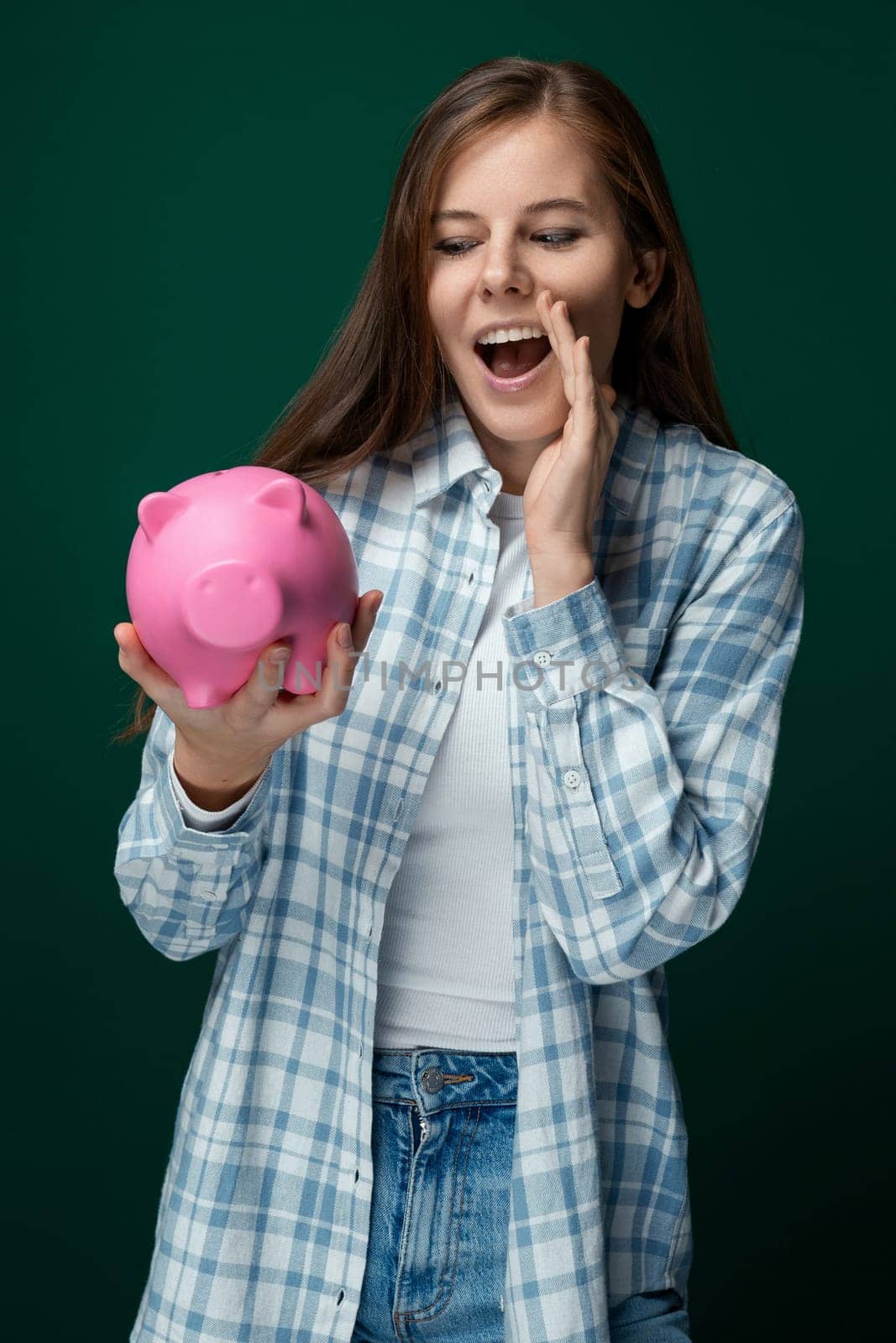 Young European woman dressed in a plaid shirt and jeans holds a piggy bank and talks about investments.