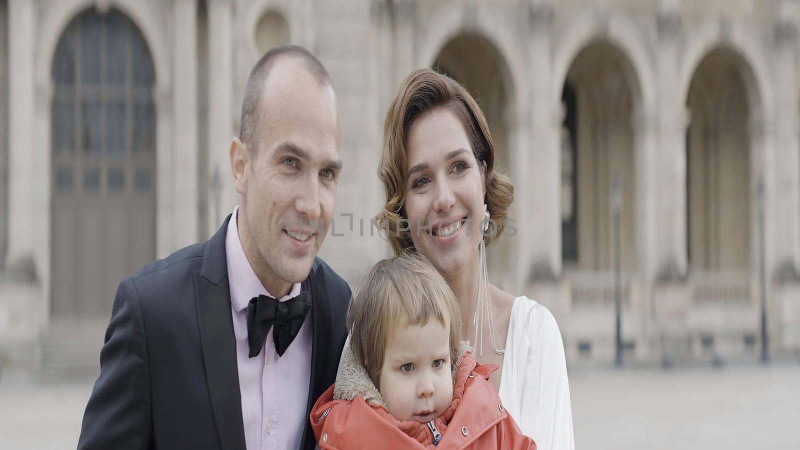 Man in suit, elegant woman in white dress, and a little girl in red jacket. Action. Happy family in a city square