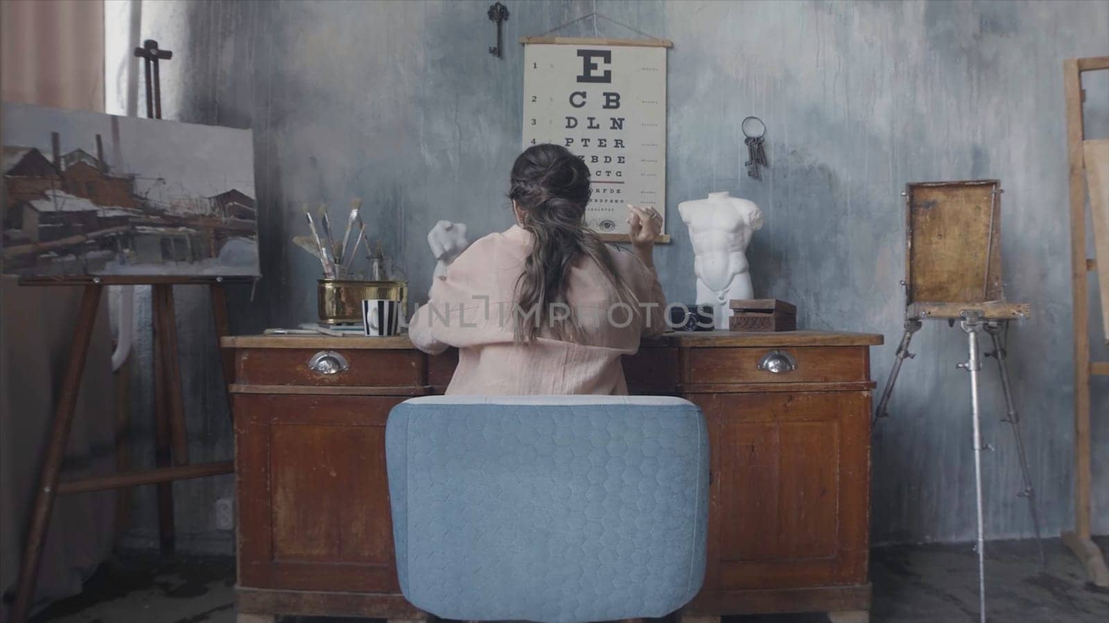 Rear view of a brunette woman sitting at her desk and touching her hair. Woman working in the art workshop on grey walls background.