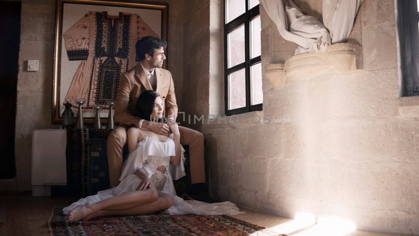 Beautiful fashionable studio shooting . Action . A beautiful young couple in wedding costumes, a bride in a magnificent dress, who are sitting on the floor in a stone room with various decorations on the walls and lighting from below. High quality 4k footage