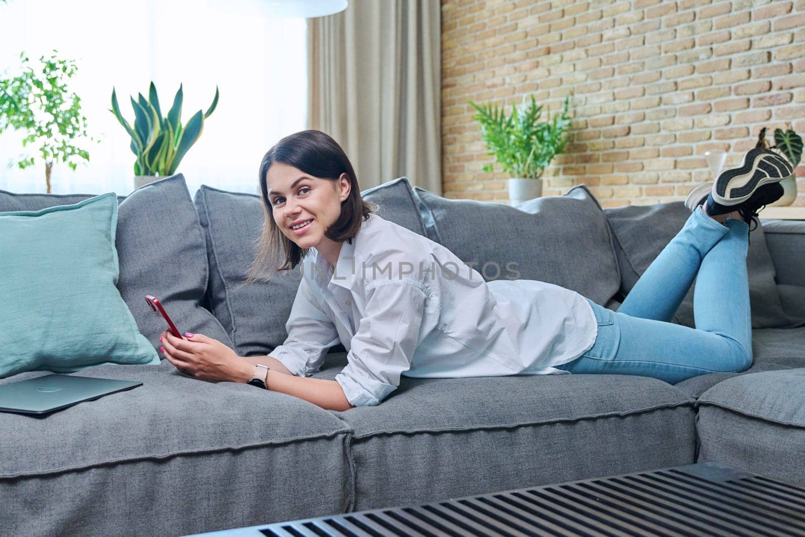 Young happy woman using smartphone, lying on sofa at home by VH-studio
