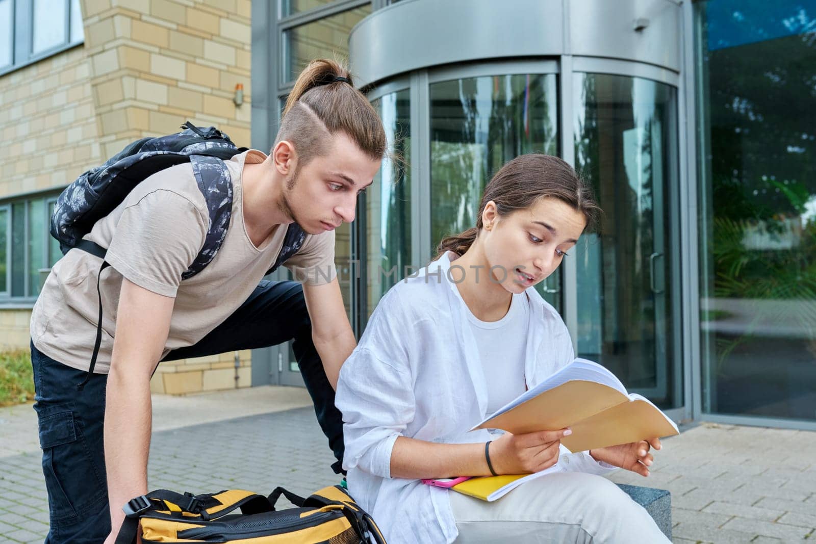 Two high school students guy and girl outdoor, school building background. Education, friendship, youth, adolescence concept