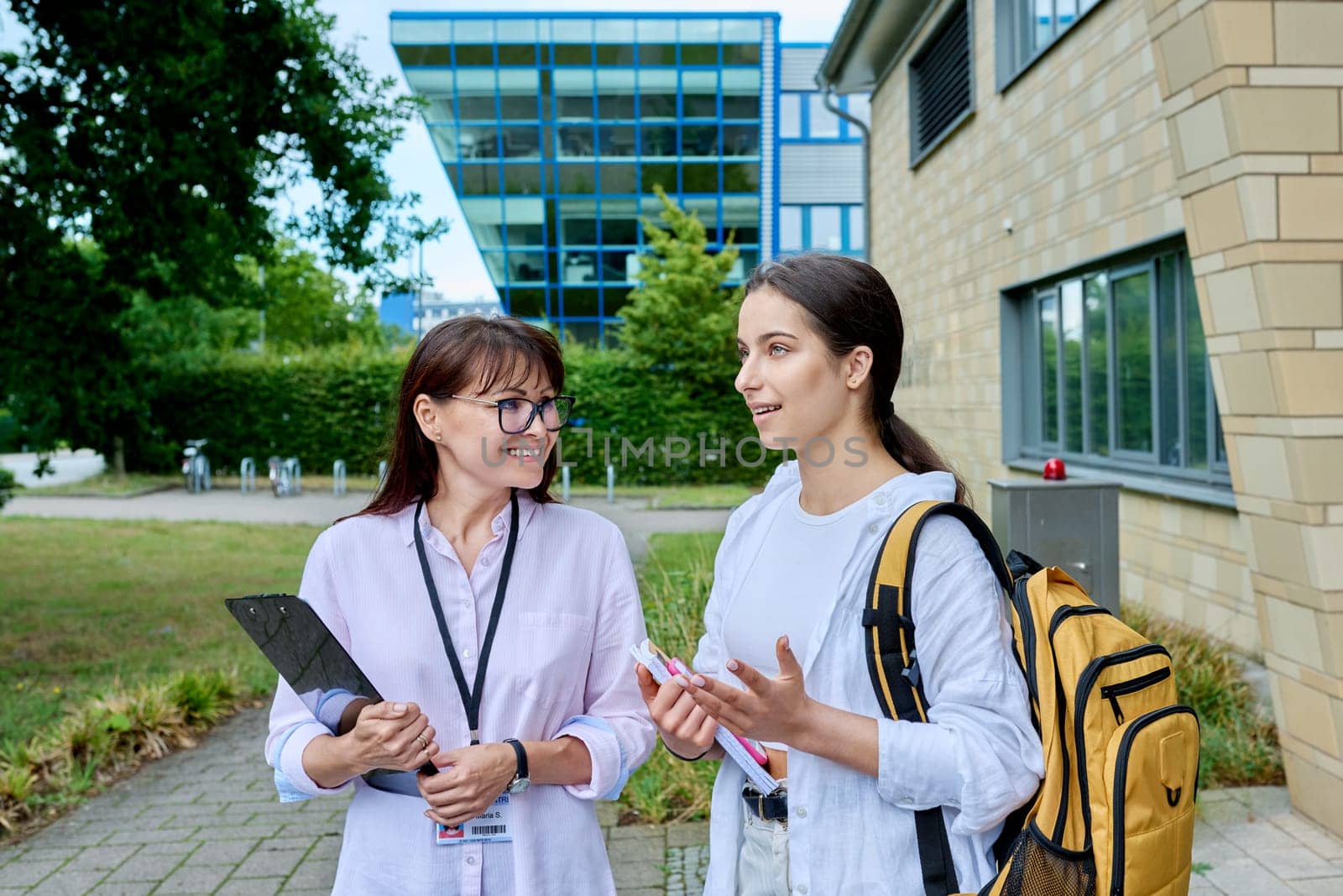 Teenage girl high school student with backpack talking to female teacher, mentor, coach, standing outdoors on educational building background. Adolescence, education, knowledge, communication