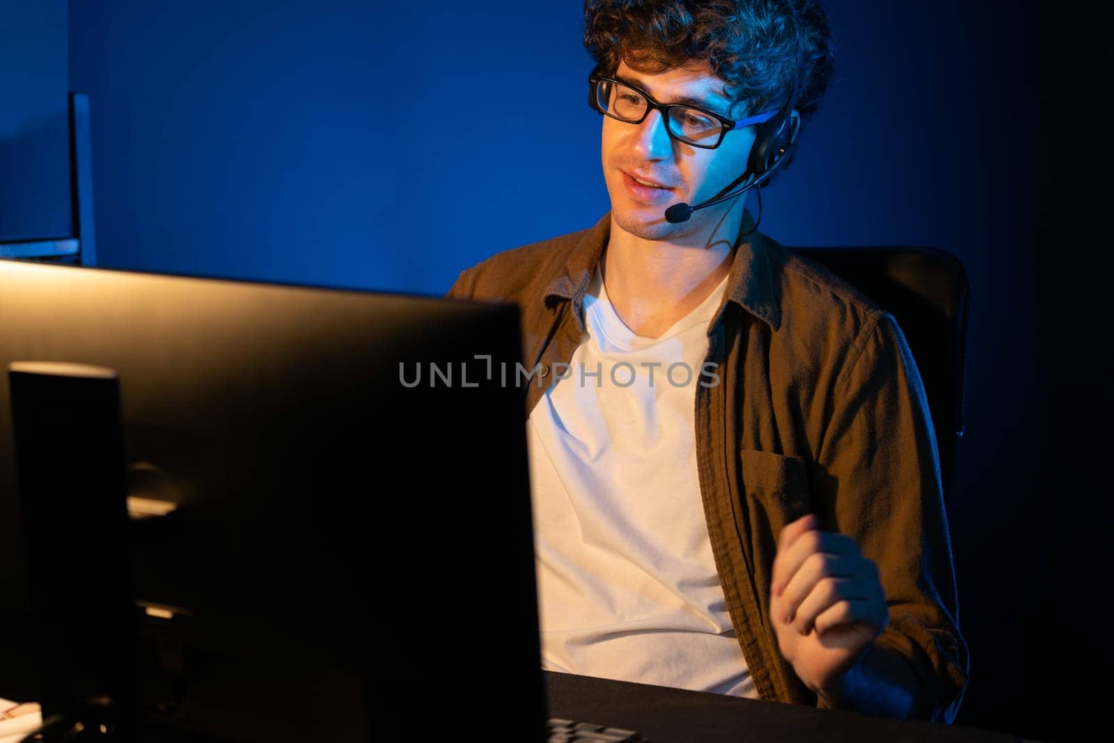 Young creative businessman communicating with listeners on laptop screen or meeting with team member, wearing headphones at neon light modern home office. Concept of working at night time. Gusher.