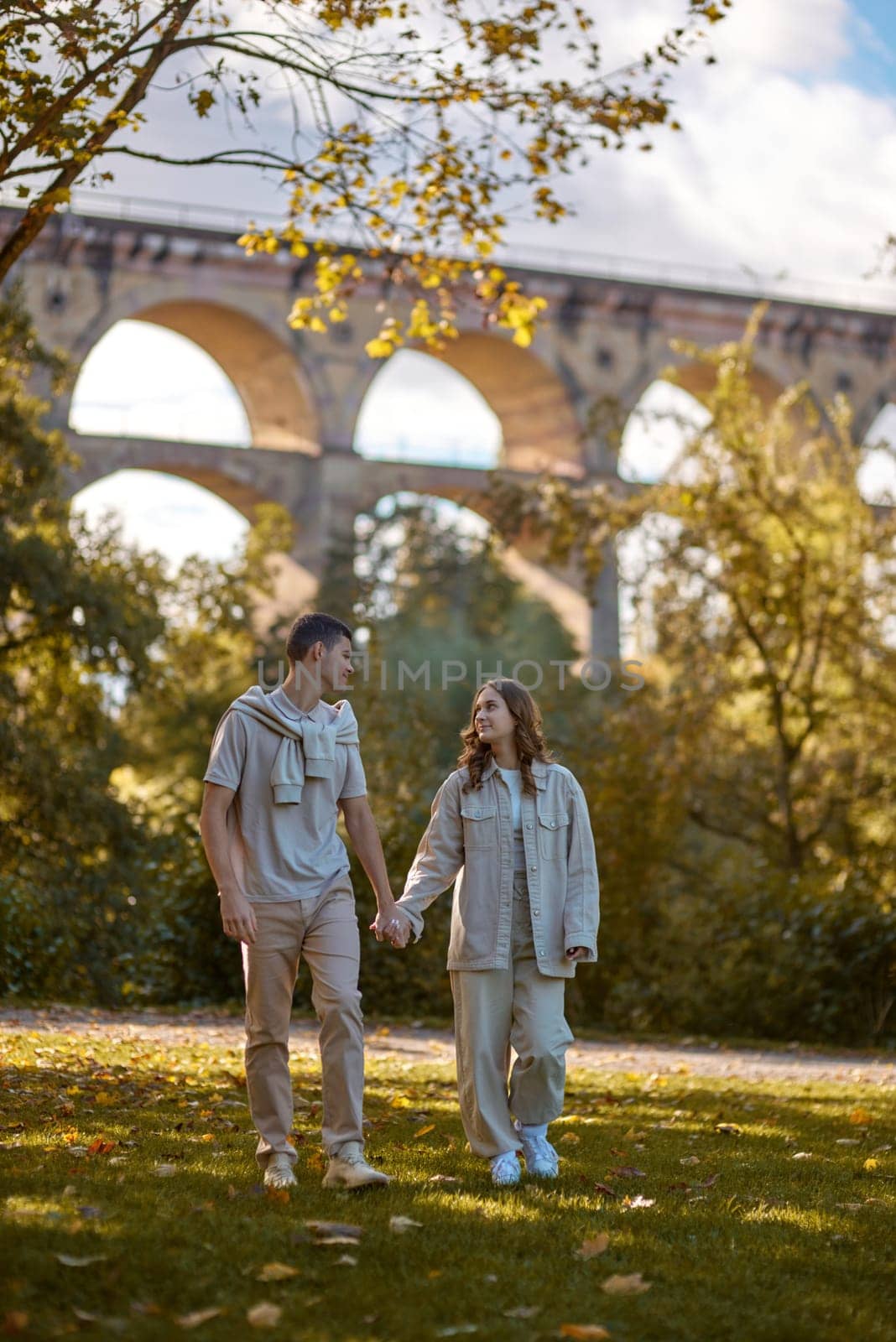 Autumn Stroll: Loving Couple Hand in Hand by Vintage Bridge in the Park. romantic escapade, autumn love, historical romance, seasonal beauty, couple goals, vintage architecture, scenic beauty, urban adventure, love story, nature exploration by Andrii_Ko