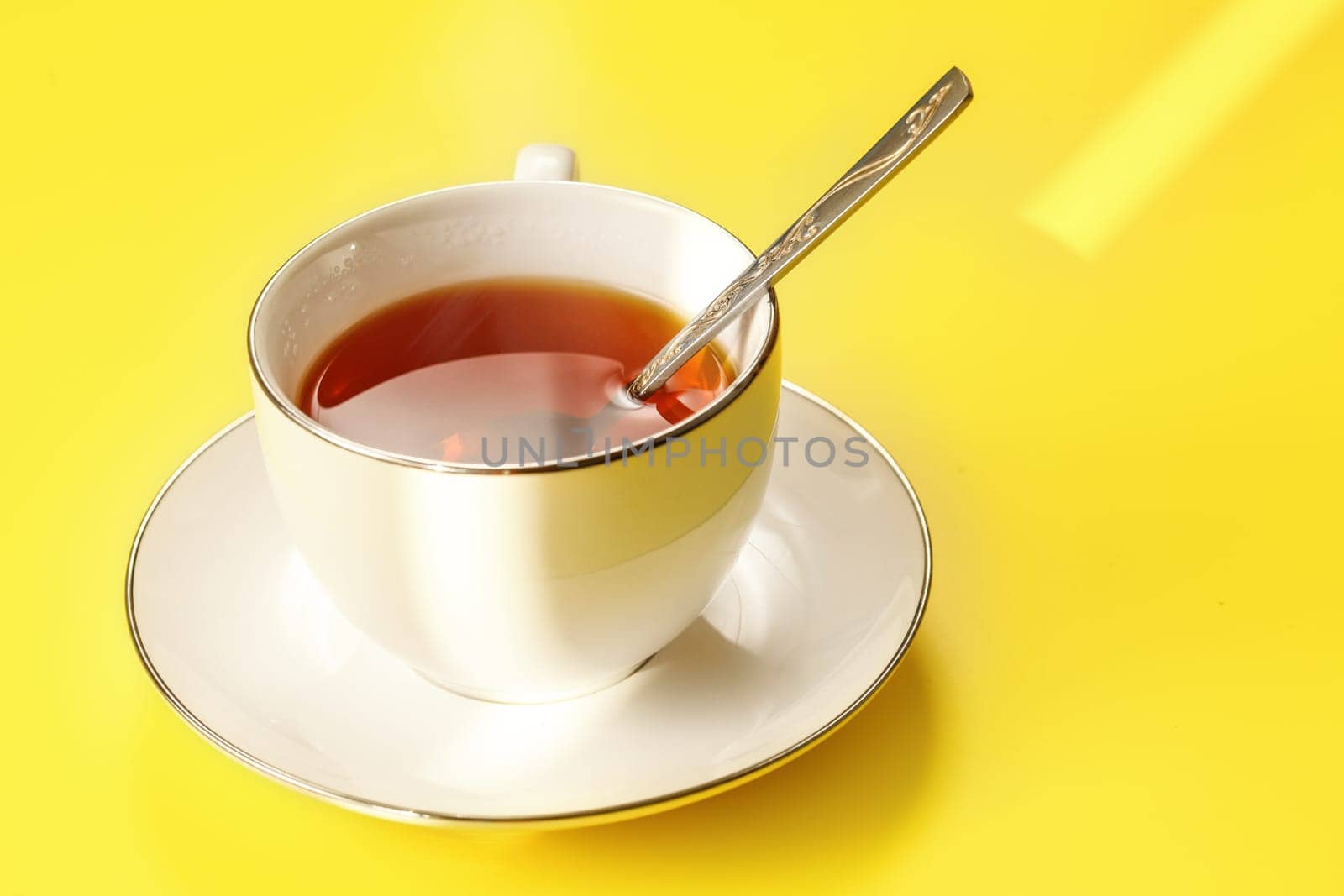 Morning sun shines to white porcelain cup with hot black tea, just brewed, silver spoon in, on yellow board by Ivanko