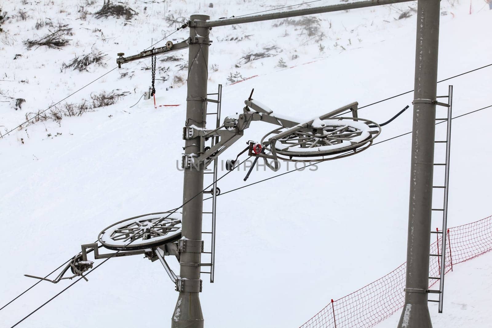 Large metal wheels for steel cables on top of ski lift supporting pillar, snow covered piste in background by Ivanko