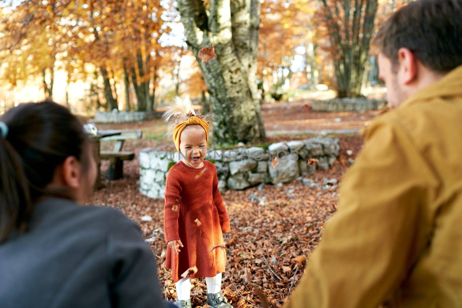 Little laughing girl under falling leaves stands in front of mom and dad in the autumn park. Back view. High quality photo