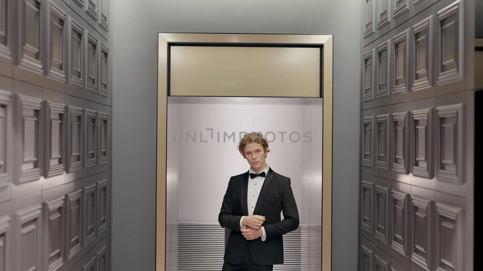 Handsome stylish groom in the hotel interior posing in front of the camera. Action. Man in black suit and white shirt standing by the wall in the end of the corridor