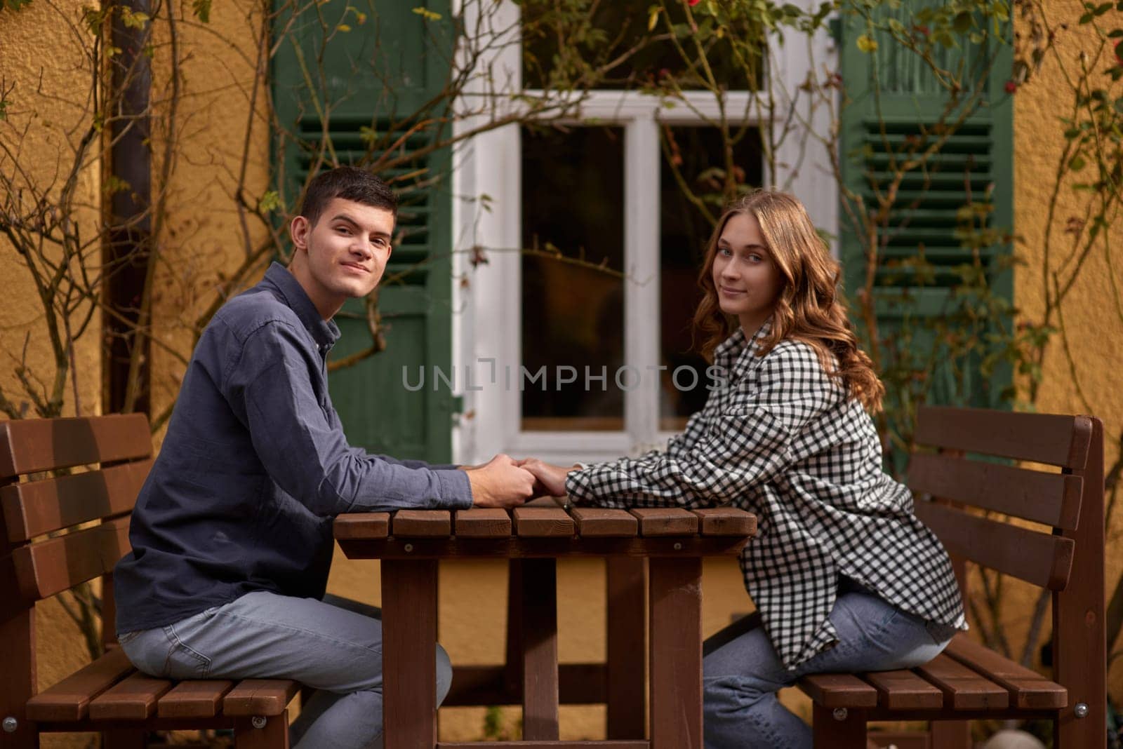 Teen Sweethearts: Holding Hands at an Outdoor Café in a European Town. Happy smiling young dating couple having coffee together and enjoying life sitting at table holding hands in street cafe on summer day. Pretty man and woman spending time together enjoying weekend. Dating, young fashion elegant stylish couple sitting at a table in a cafe , gently hold hands, on streets of old european city. American beautiful man hugging caucasian woman and sitting at cafe, hat on table. Concept of happy international c by Andrii_Ko
