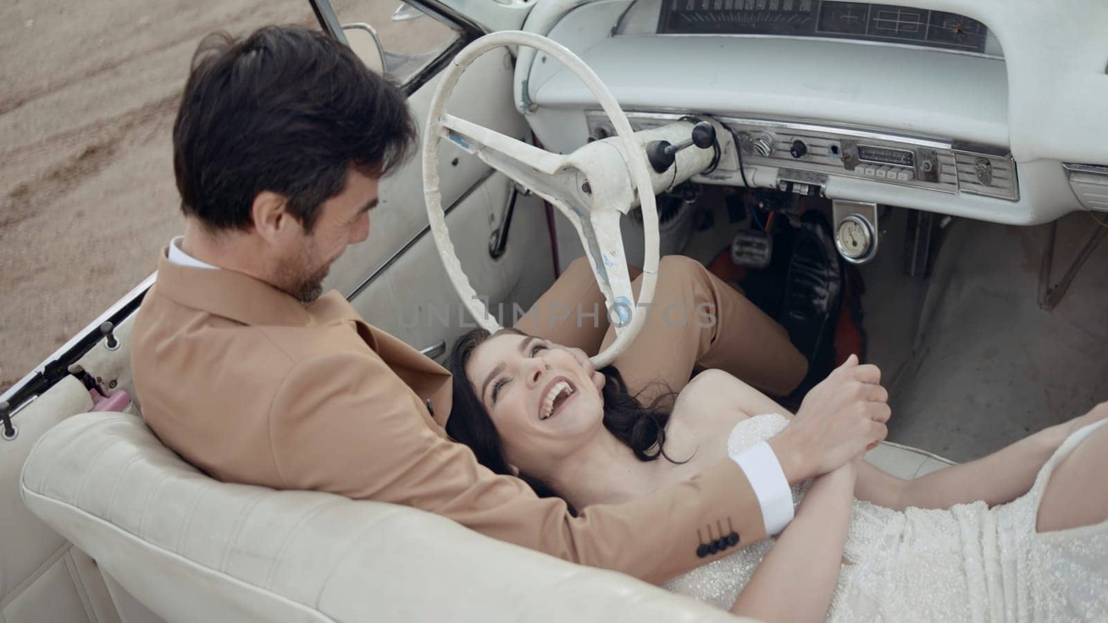 Happy smiling woman lying on the knees of her husband inside the cabriolet car. Action. Groom in suit holding hand of a beautiful bride