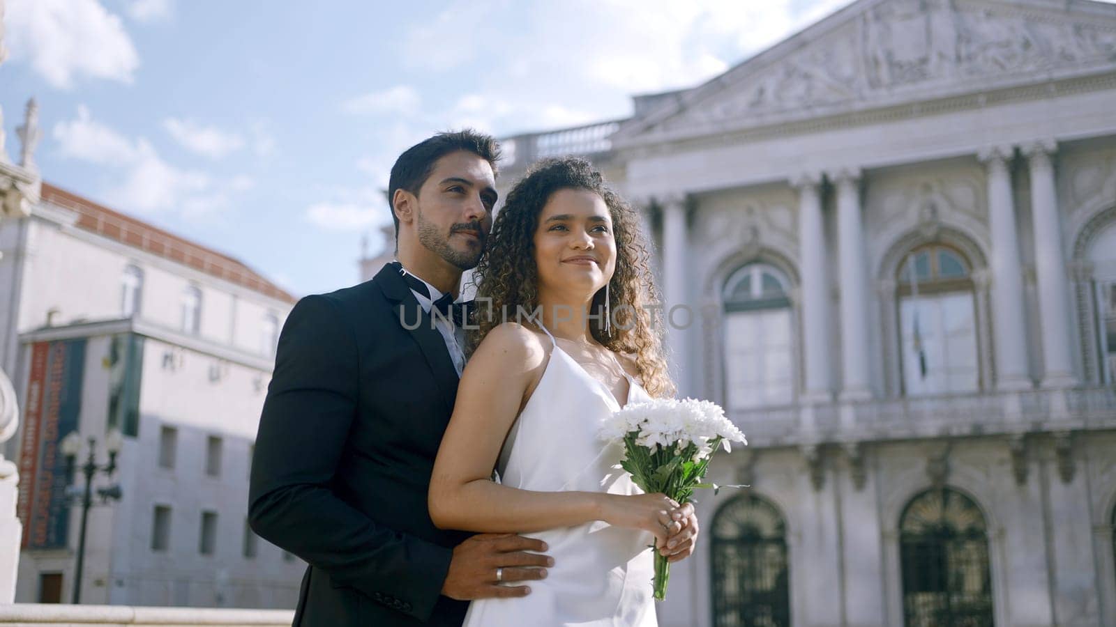 Elegant couple happily hugging. Action. Stylish couple in luxurious outfits hugging in city center. Newlyweds hug on background of ancient buildings on sunny summer day.