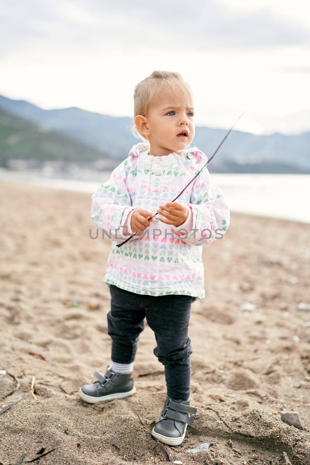 Little girl stands on the beach with a stick in her hands and looks into the distance by Nadtochiy