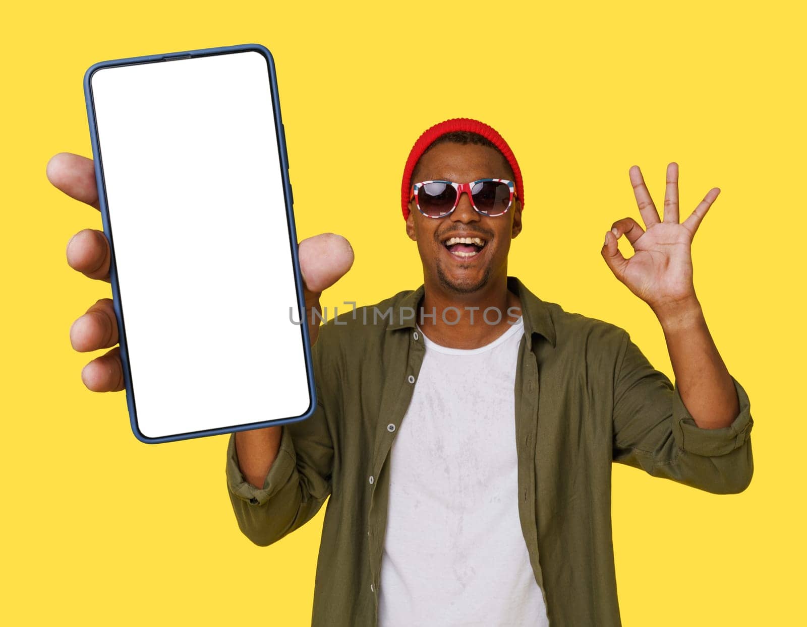 Trendy Young Man In Casual Closing Shows Ok Or Okay Gesture With Mobile Phone With Empty White Screen For Copy Space Isolated On Yellow Background. High quality photo