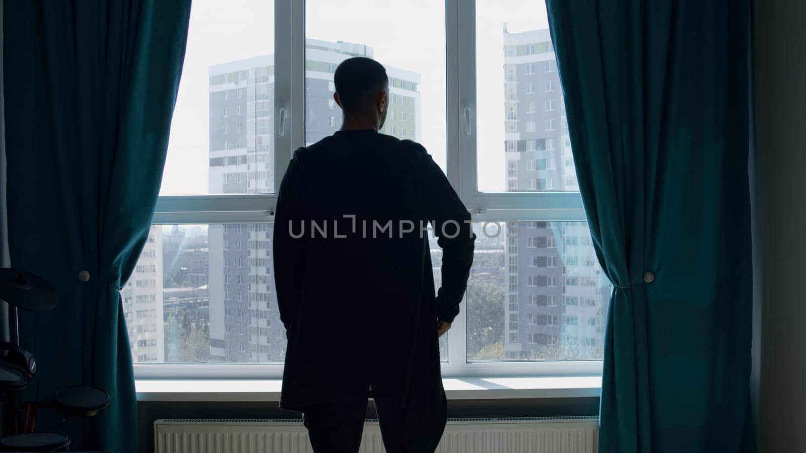 Rear view of a dressed man standing indoors in a living room by the window. Media. Silhouette of a man looking through the window