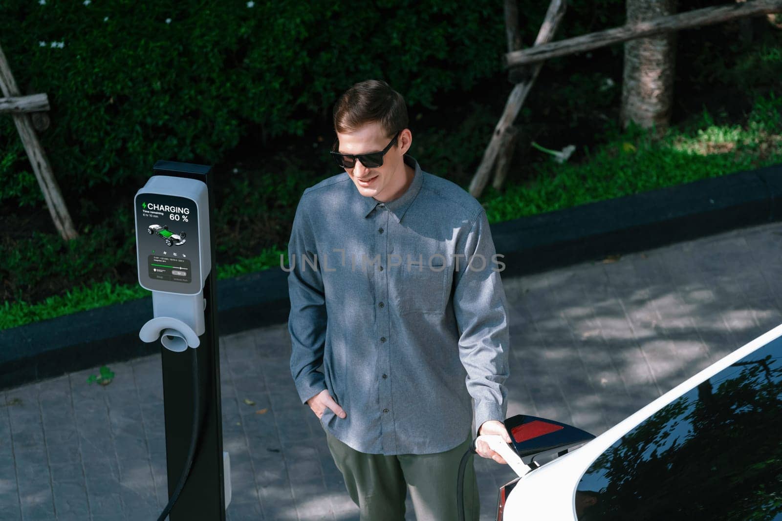 Young man travel with EV electric car charging in green sustainable city outdoor garden in summer shows urban sustainability lifestyle by green clean rechargeable energy of electric vehicle innards