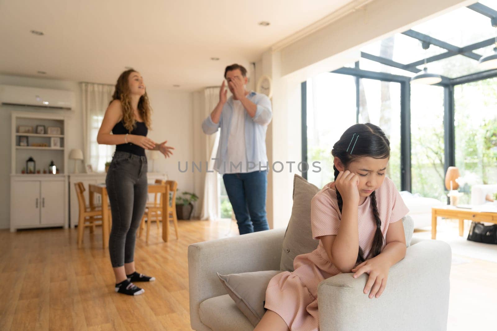 Annoyed and unhappy young girl sitting on sofa trapped in middle of tension by her parent argument in living room. Unhealthy domestic lifestyle and traumatic childhood develop to depression.Synchronos