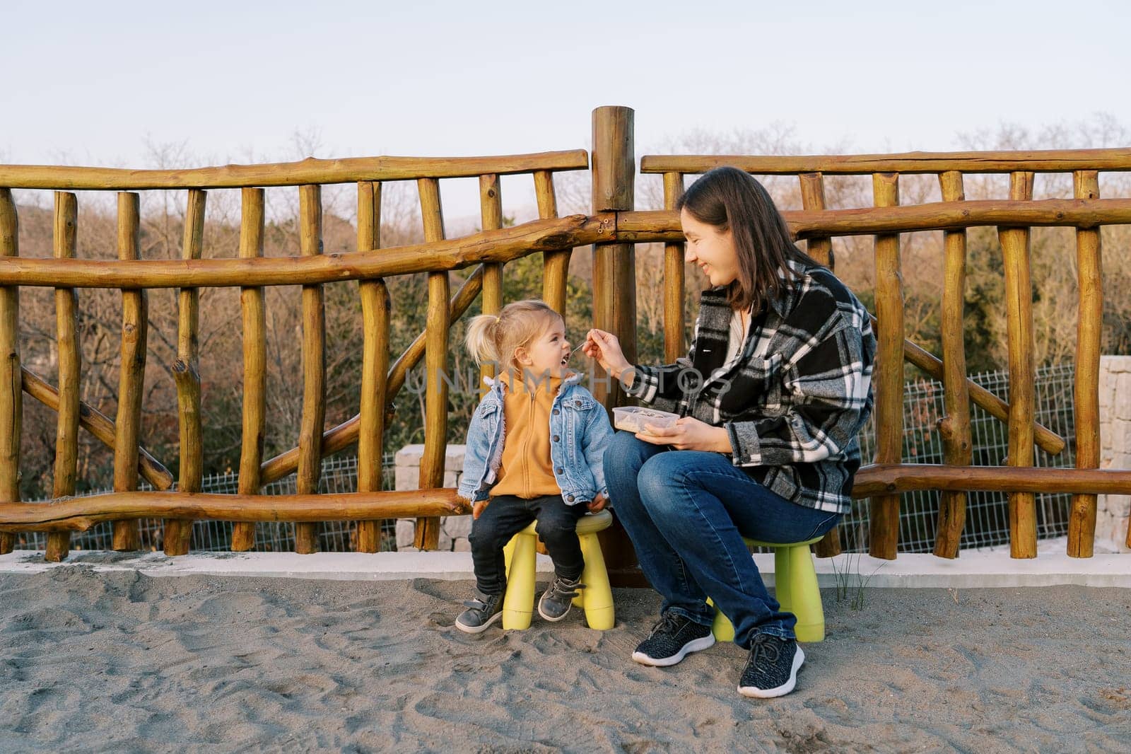 Mom feeds a little girl from a spoon while sitting on chairs near a wooden fence. High quality photo