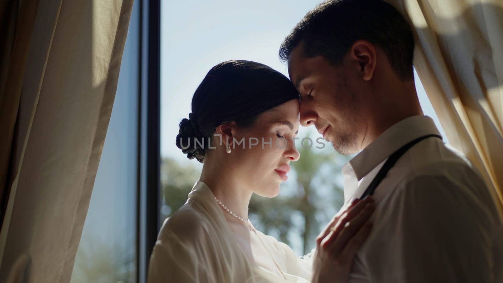 Young elegant couple hugging at the window. Action. A beautiful woman in a white suit with a man on whom the sun's rays fall. by Mediawhalestock