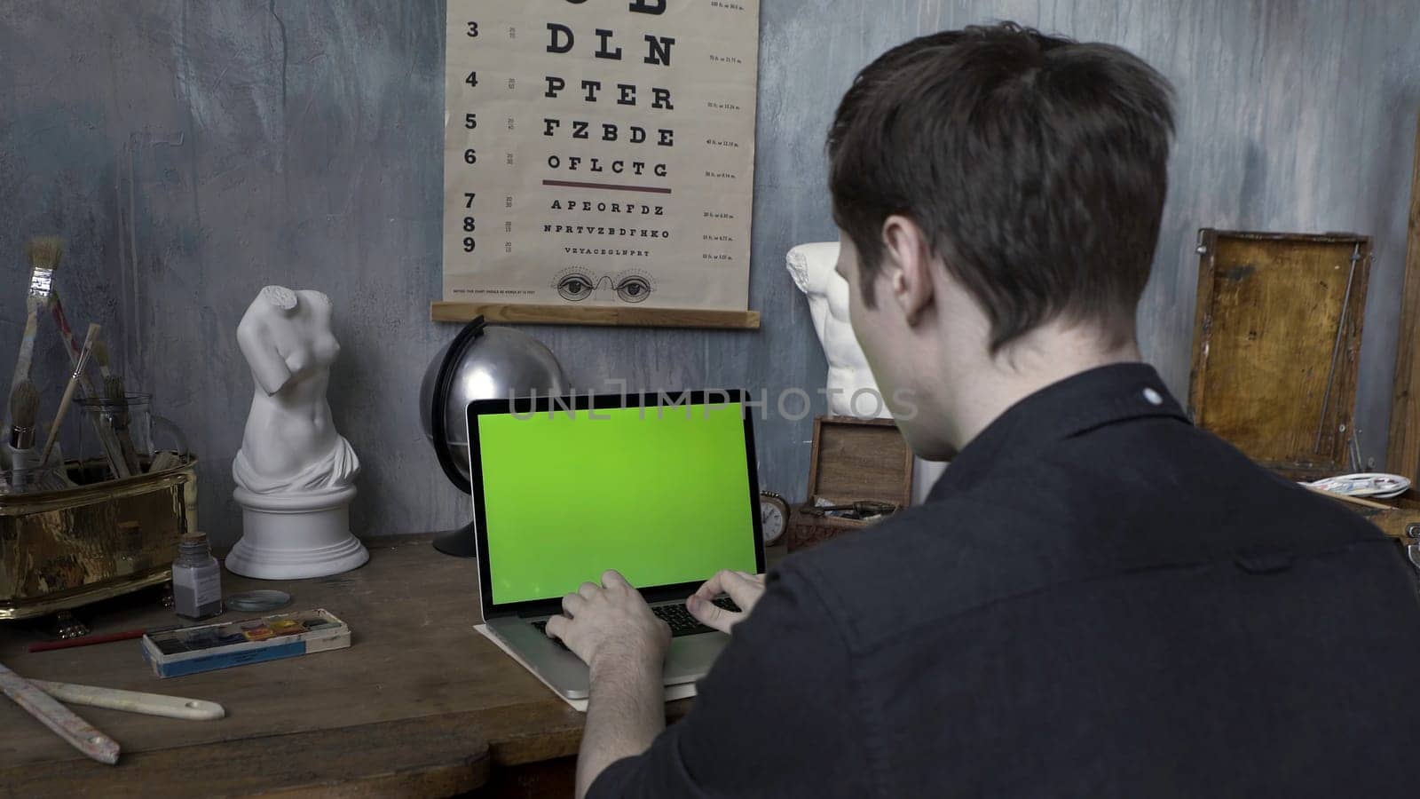 Rear view of the male artisan working at desk on laptop with chromakey in the artist workshop. Man typing on the computer with green screen in the decorated room.