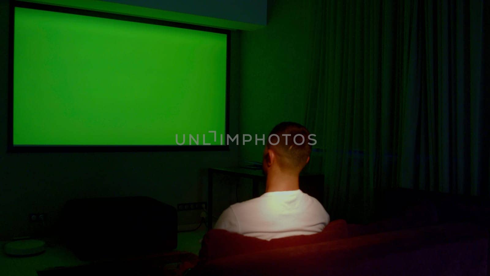 Man watching TV with green screen in living room, back view. Media. Man switching channels with hand gestures, gesture interface, modern technology